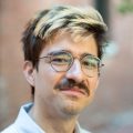 A headshot of reporter Eddie Velazquez wearing a blue button up shirt and glasses. He has a moustache and brown hair with blonde streaks.